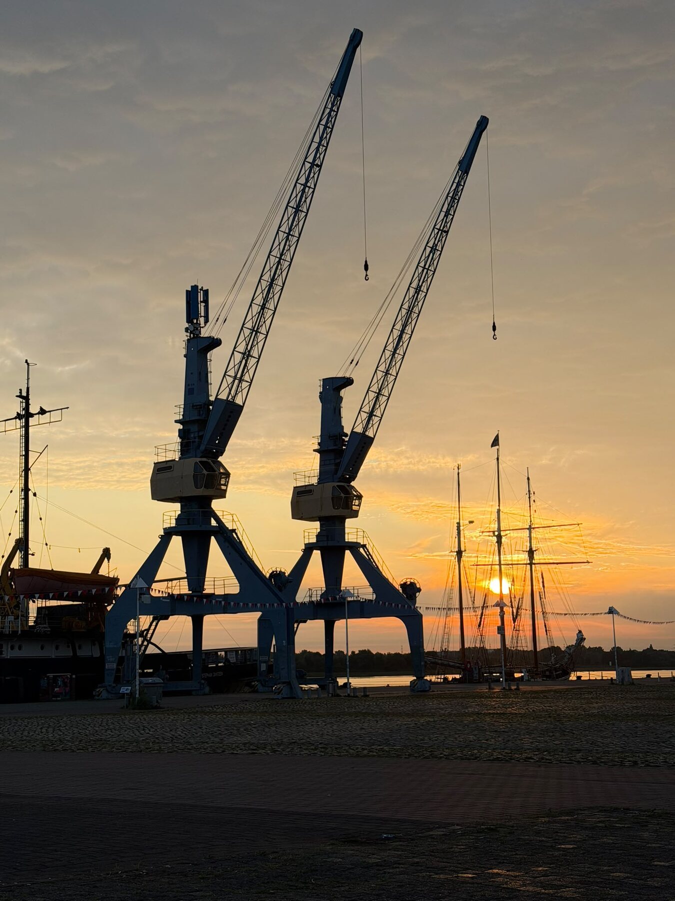Zwei Hafenkräne und ein Segelschiff vor einem goldenen Sonnenuntergang